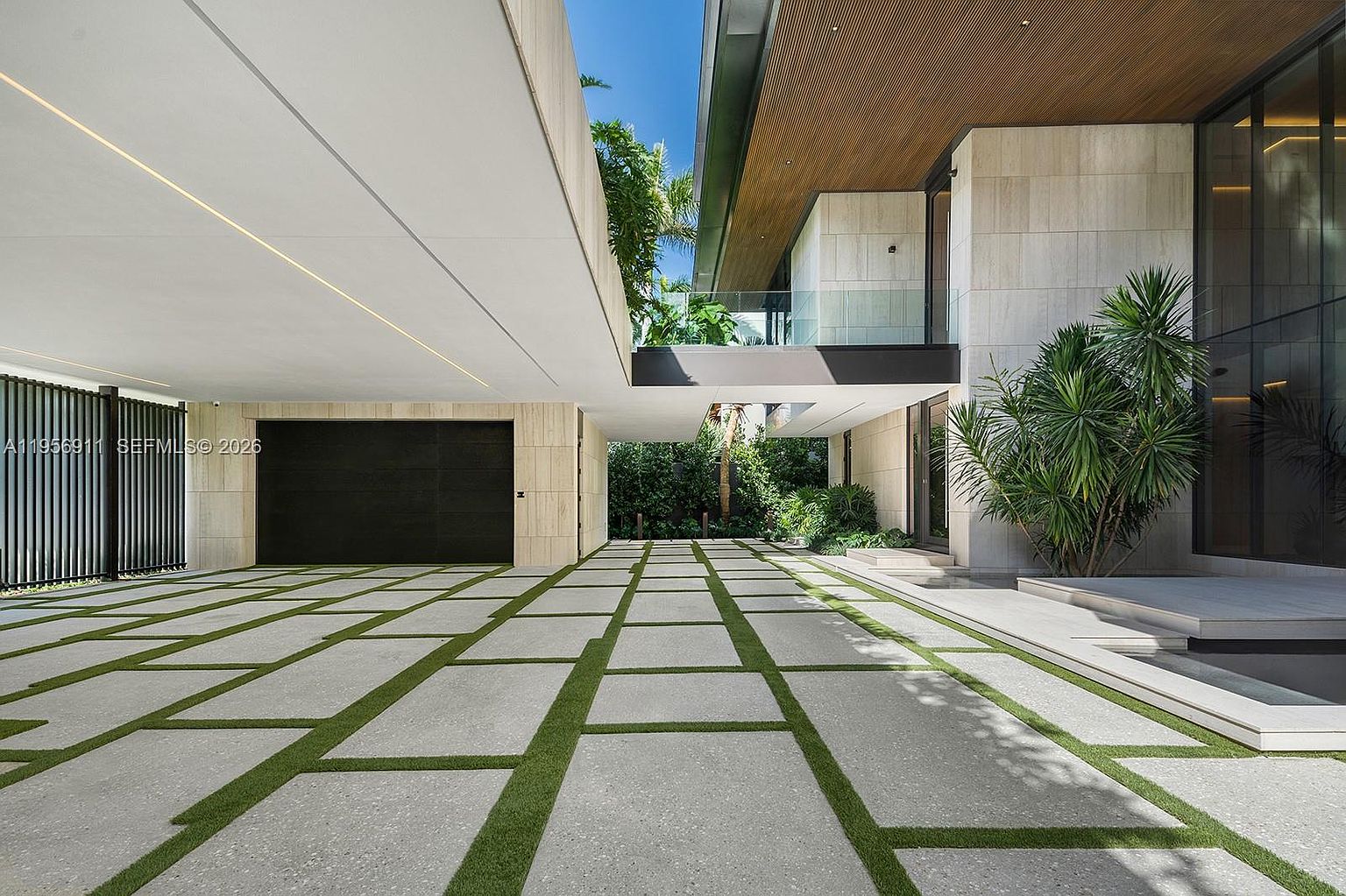 This modern architectural exterior features a spacious covered driveway leading to a sleek, dark-toned garage door. The ground is paved with large, light-colored stone pavers separated by strips of lush green grass, creating a geometric pattern that leads toward the home's entrance. The structure showcases clean lines, stone-clad walls, and a prominent wood-slatted ceiling overhang, emphasizing a sophisticated and contemporary aesthetic.