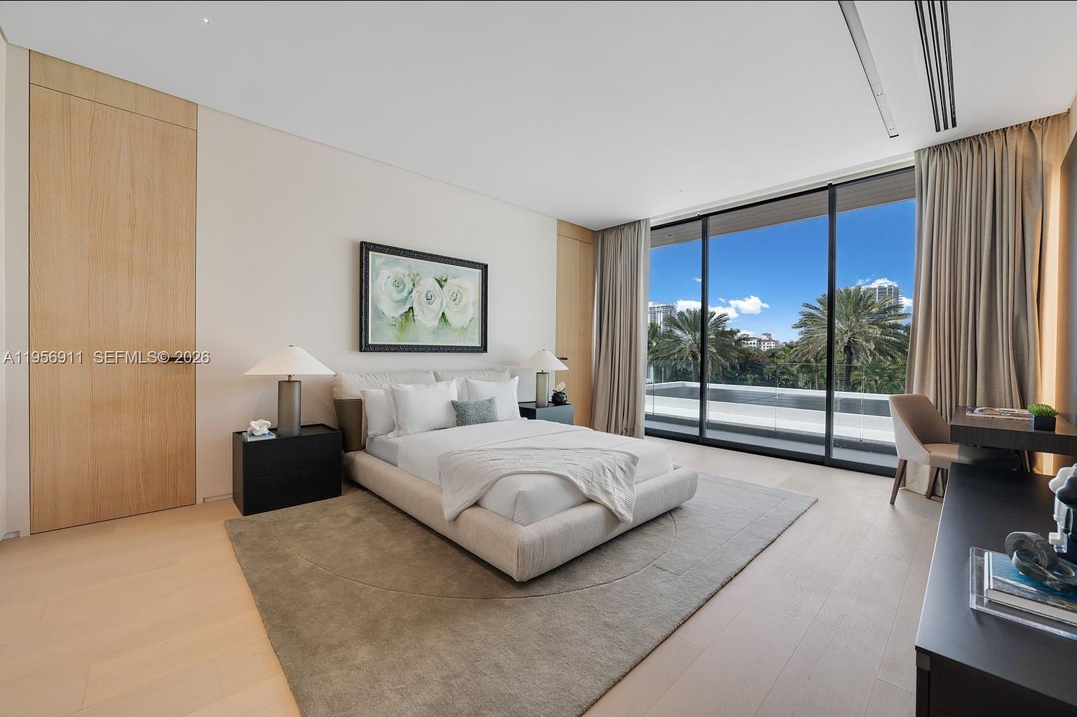 This spacious primary bedroom features a minimalist aesthetic with light wood flooring and a large area rug anchoring the bed. A wall of floor-to-ceiling windows provides abundant natural light and a view of an outdoor terrace with a pool, while a dedicated desk area is positioned to the side. The room is decorated with neutral tones and a large floral artwork above the bed, creating a serene and sophisticated atmosphere.