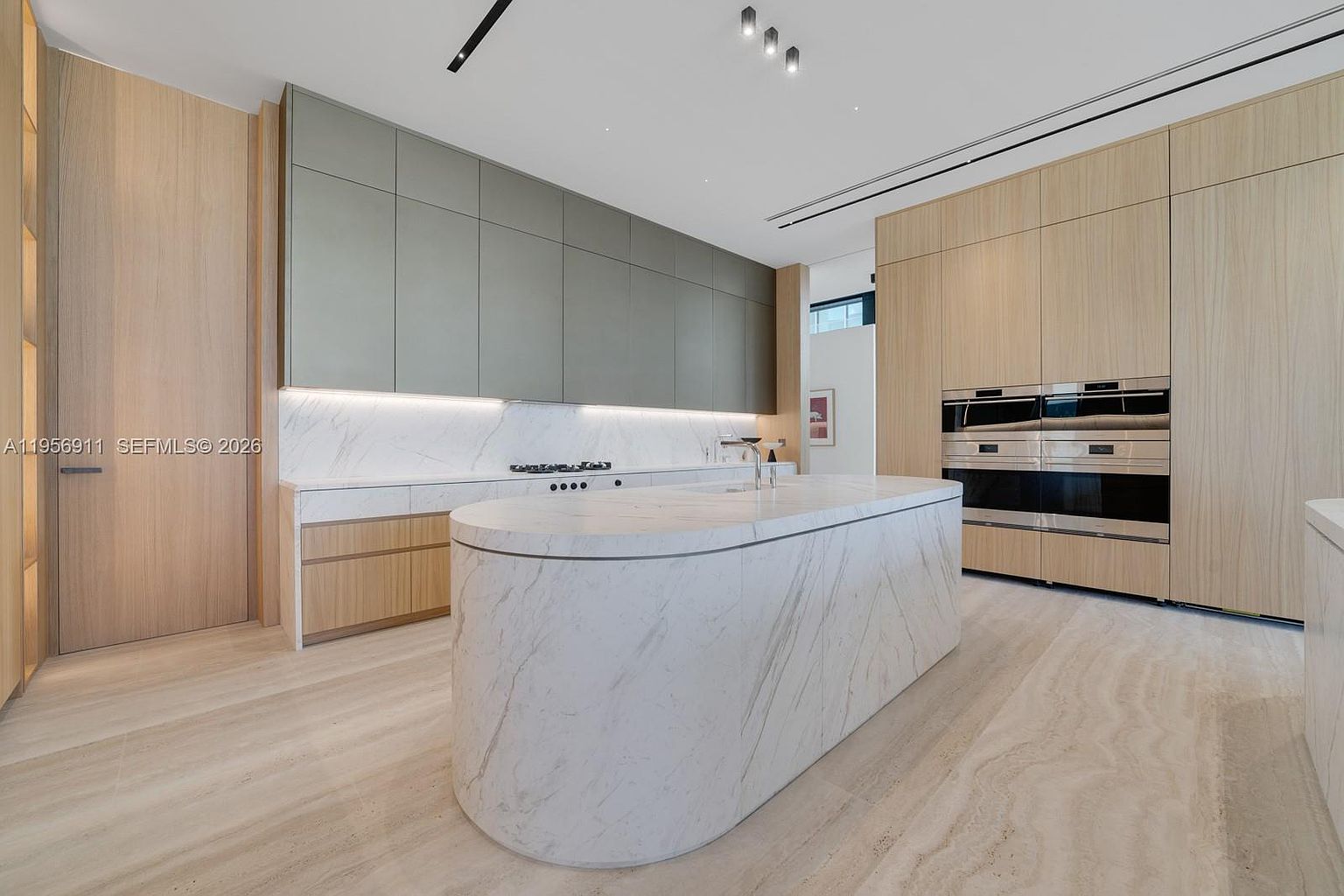 This modern, minimalist kitchen features a large, curved stone island as its centerpiece, complemented by light wood cabinetry and sleek, matte-finish upper cabinets. The space is illuminated by recessed lighting and under-cabinet accent lights, highlighting the clean lines and integrated appliances. The perspective is a wide-angle shot from the kitchen floor, showcasing the seamless transition between the stone surfaces and the light-toned flooring.