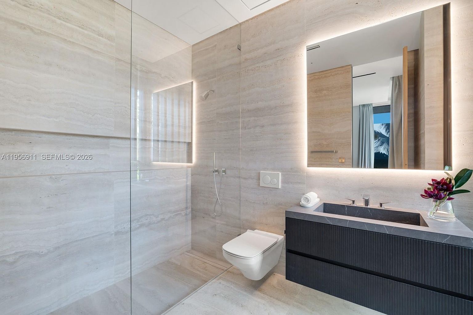 This modern, minimalist bathroom features large-format stone wall tiles and a sleek, wall-mounted vanity with a dark, textured finish. A glass partition separates the walk-in shower area from the rest of the room, which includes a wall-mounted toilet and a large, backlit mirror. The space is characterized by clean lines, neutral tones, and a sophisticated, contemporary aesthetic.