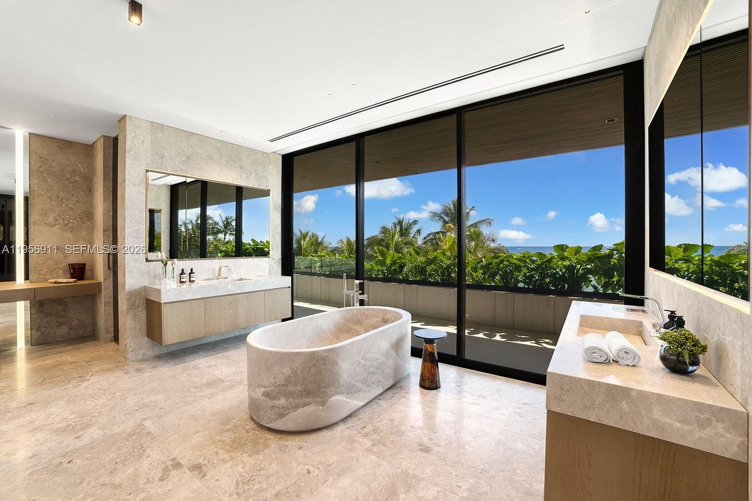 This luxurious primary bathroom features a central stone soaking tub and dual vanities with light wood cabinetry and stone countertops. Large, floor-to-ceiling windows provide an expansive view of the ocean and lush greenery, filling the space with natural light. The room is finished with neutral stone flooring and walls, creating a serene and high-end spa-like atmosphere.