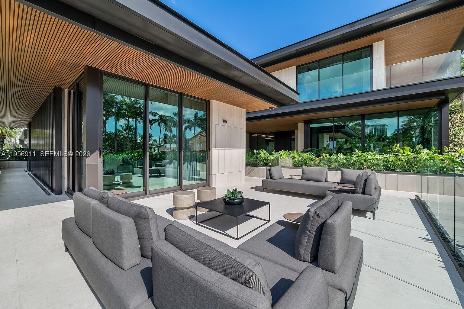 This expansive, modern outdoor terrace features sleek gray sectional seating arranged around a central black coffee table, creating a sophisticated lounge area. The space is defined by clean lines, large glass sliding doors, and a wood-slatted ceiling overhang that provides architectural interest. Lush greenery lines the perimeter, and the open-air design offers a seamless transition between indoor and outdoor living spaces.