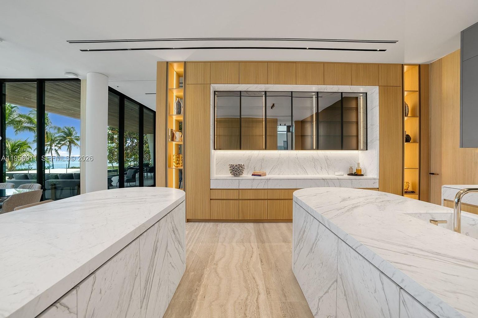 This sophisticated, modern kitchen features dual islands with light-toned stone surfaces and a matching backsplash. The cabinetry is crafted from light wood, complemented by integrated shelving and a central mirrored display area. Large glass panels provide a seamless view of the lush outdoor greenery, creating a bright and airy atmosphere.