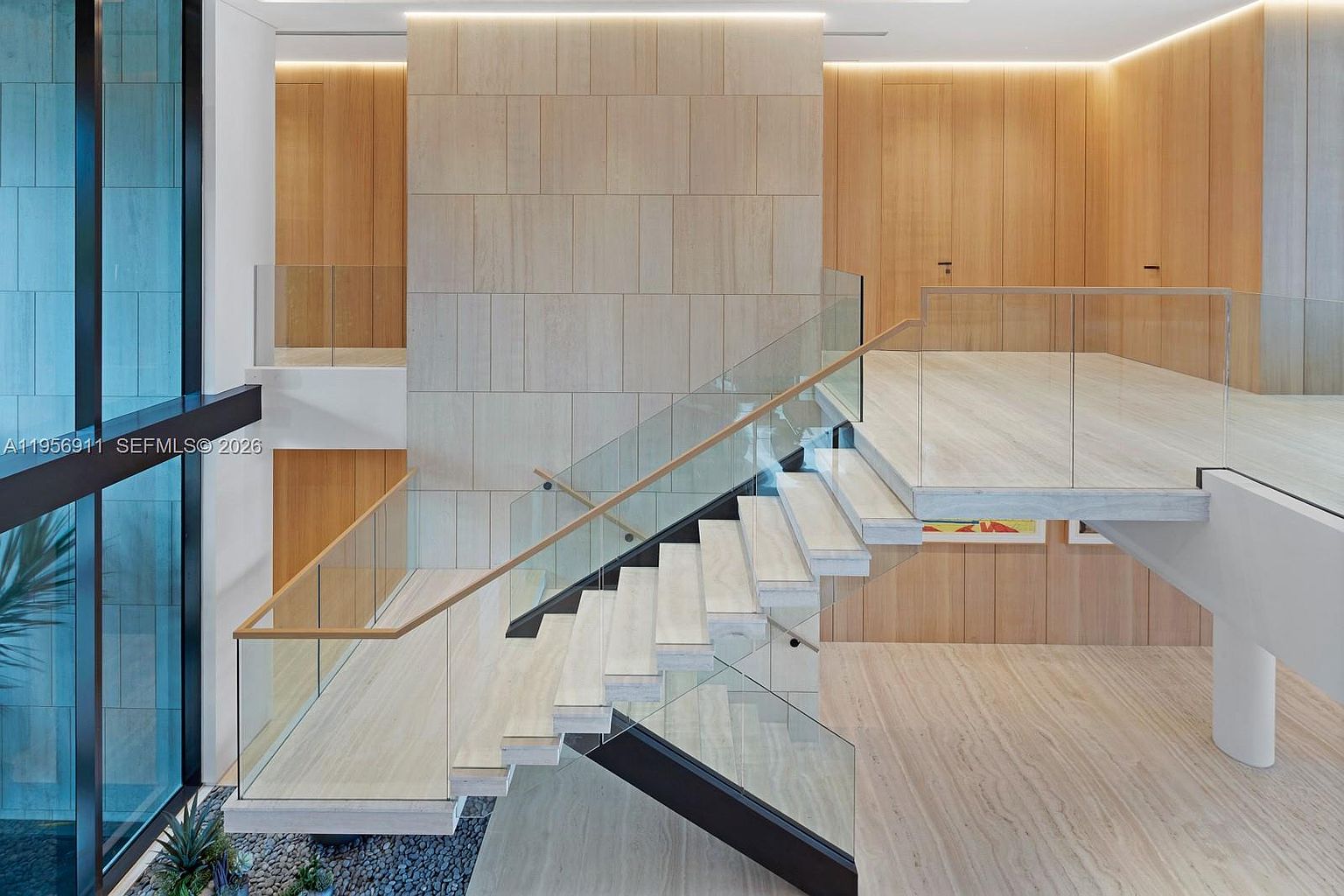 This striking interior space features a modern floating staircase with glass railings and light wood treads, creating an open and airy aesthetic. A central wall clad in large, light-toned stone panels serves as a focal point, complemented by warm wood paneling and integrated cove lighting. The perspective looks down from a landing, highlighting the architectural geometry and the contrast between the sleek glass, natural stone, and wood textures.