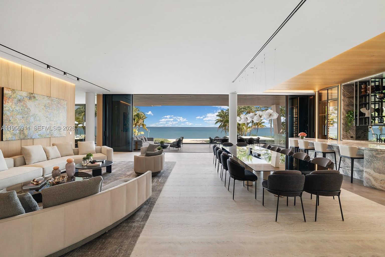 This expansive, open-concept living and dining area features a seamless transition to an outdoor terrace overlooking the ocean through large glass walls. The space is anchored by a neutral-toned seating area with a large abstract painting and a long dining table that seats twelve, all set against a backdrop of light-toned stone flooring and wood-paneled walls. The design emphasizes a sophisticated, airy aesthetic with high-end finishes and a direct, unobstructed view of the horizon.
