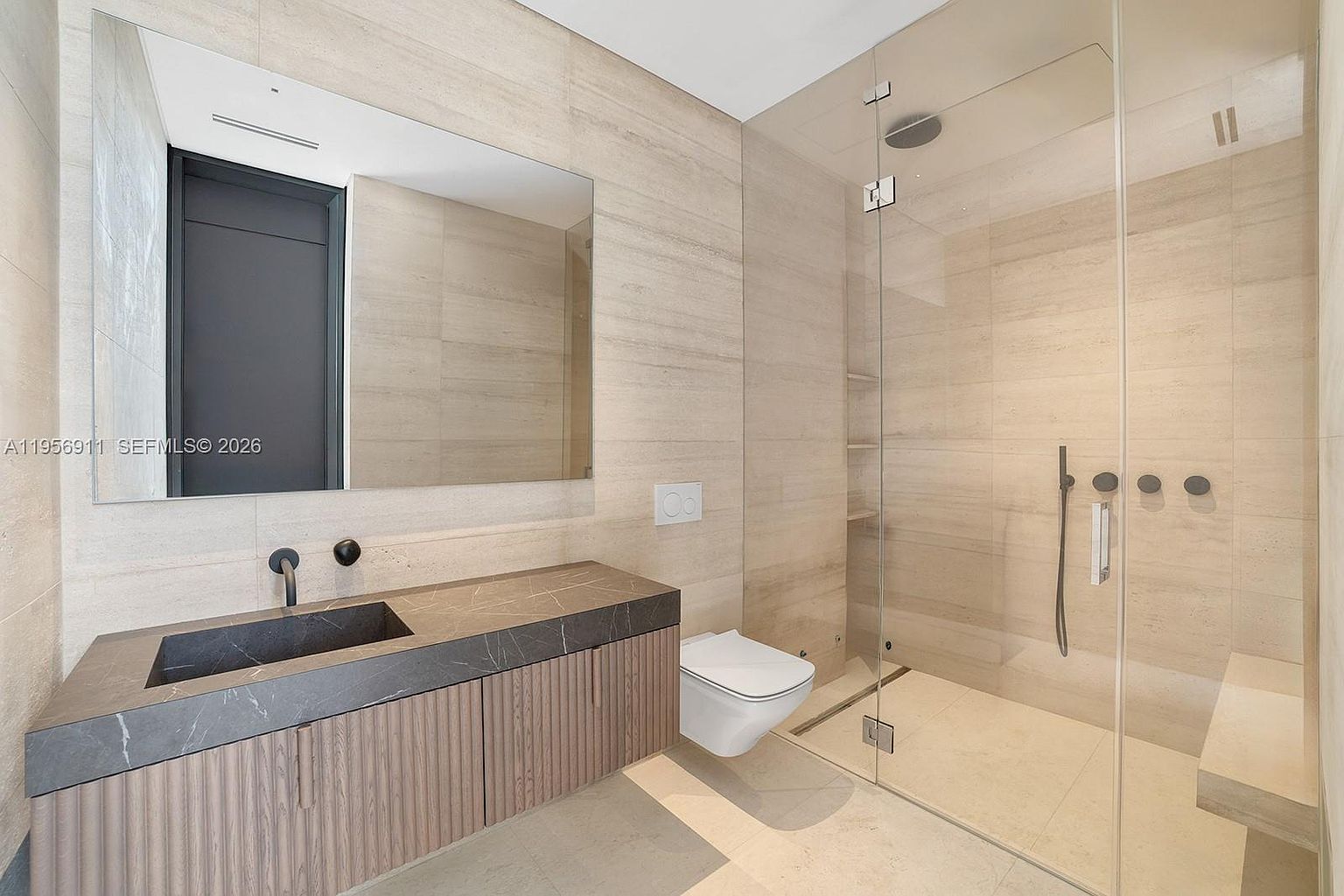 This modern bathroom features a sleek floating vanity with a dark stone countertop and a minimalist black faucet. A large, frameless glass-enclosed shower occupies the right side of the room, showcasing light-toned stone wall tiles and a built-in bench. The space is completed by a wall-mounted toilet and a large, expansive mirror that enhances the bright, contemporary aesthetic.