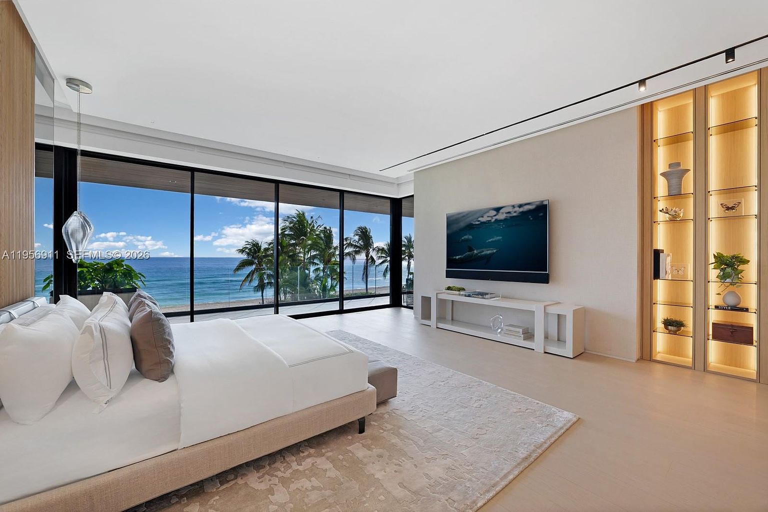 This luxurious primary bedroom features a minimalist aesthetic with light-toned flooring and a neutral color palette. A large bed faces a wall-mounted television and a low-profile media console, while floor-to-ceiling windows provide an expansive, unobstructed view of the ocean and palm trees. To the right, a built-in, illuminated display shelving unit adds a sophisticated touch to the serene and airy space.