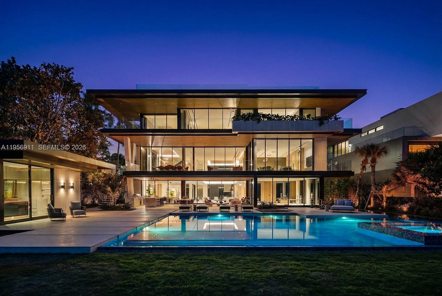 This stunning contemporary residence is captured at twilight, showcasing a multi-level architectural design with extensive glass walls that provide a seamless transition between indoor and outdoor living spaces. The property features a large, illuminated swimming pool set within a manicured lawn, complemented by a spacious patio area perfect for evening entertaining. The structure's clean lines, warm wood-toned soffits, and modern lighting create a sophisticated and luxurious atmosphere.