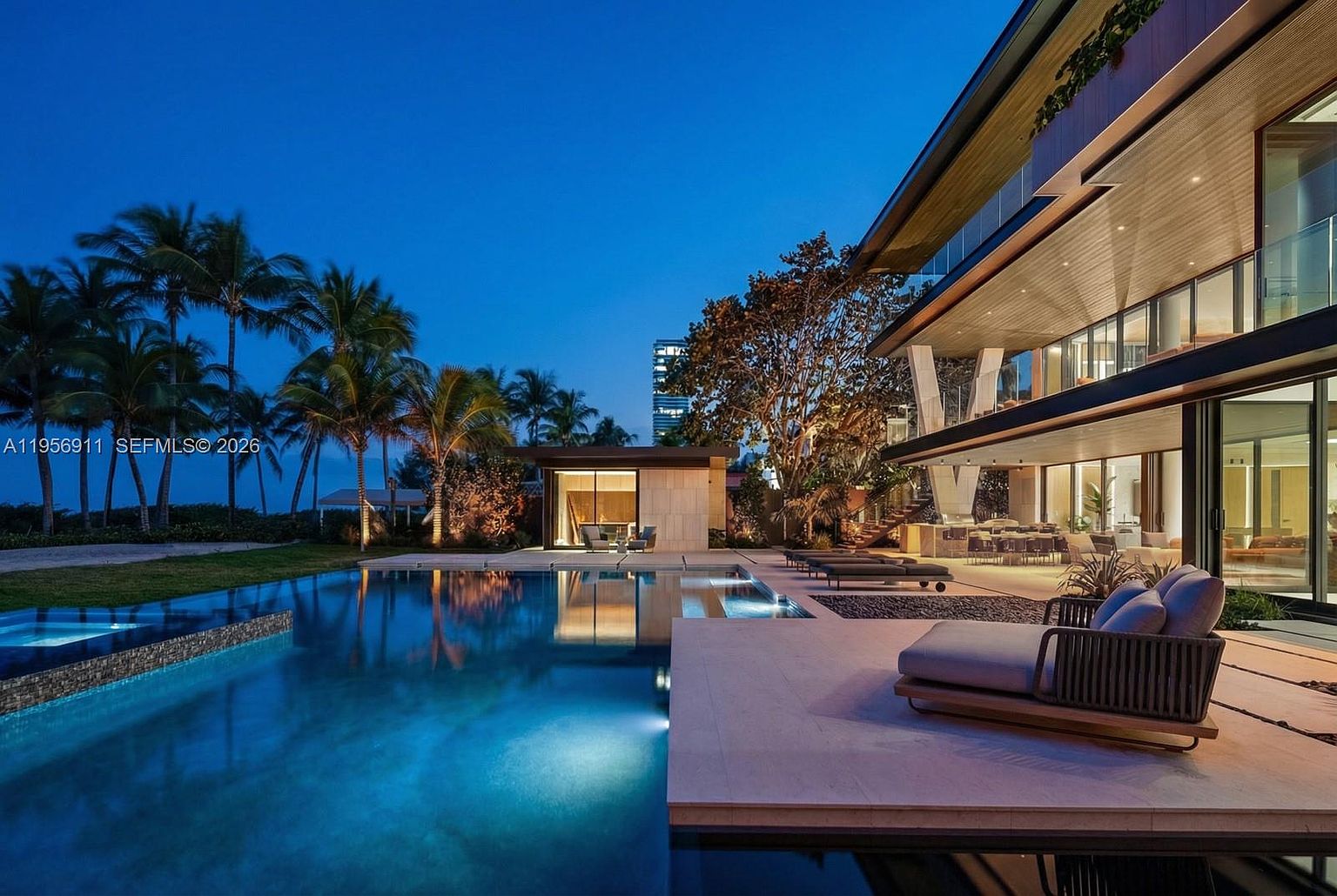 This stunning twilight view captures a modern luxury residence with a sprawling infinity-edge pool that reflects the deep blue evening sky. The expansive stone deck features contemporary lounge chairs and a seamless transition between the indoor living areas and the outdoor space, highlighted by warm ambient lighting. Palm trees and lush landscaping frame the scene, creating a serene and sophisticated atmosphere perfect for high-end outdoor entertaining.
