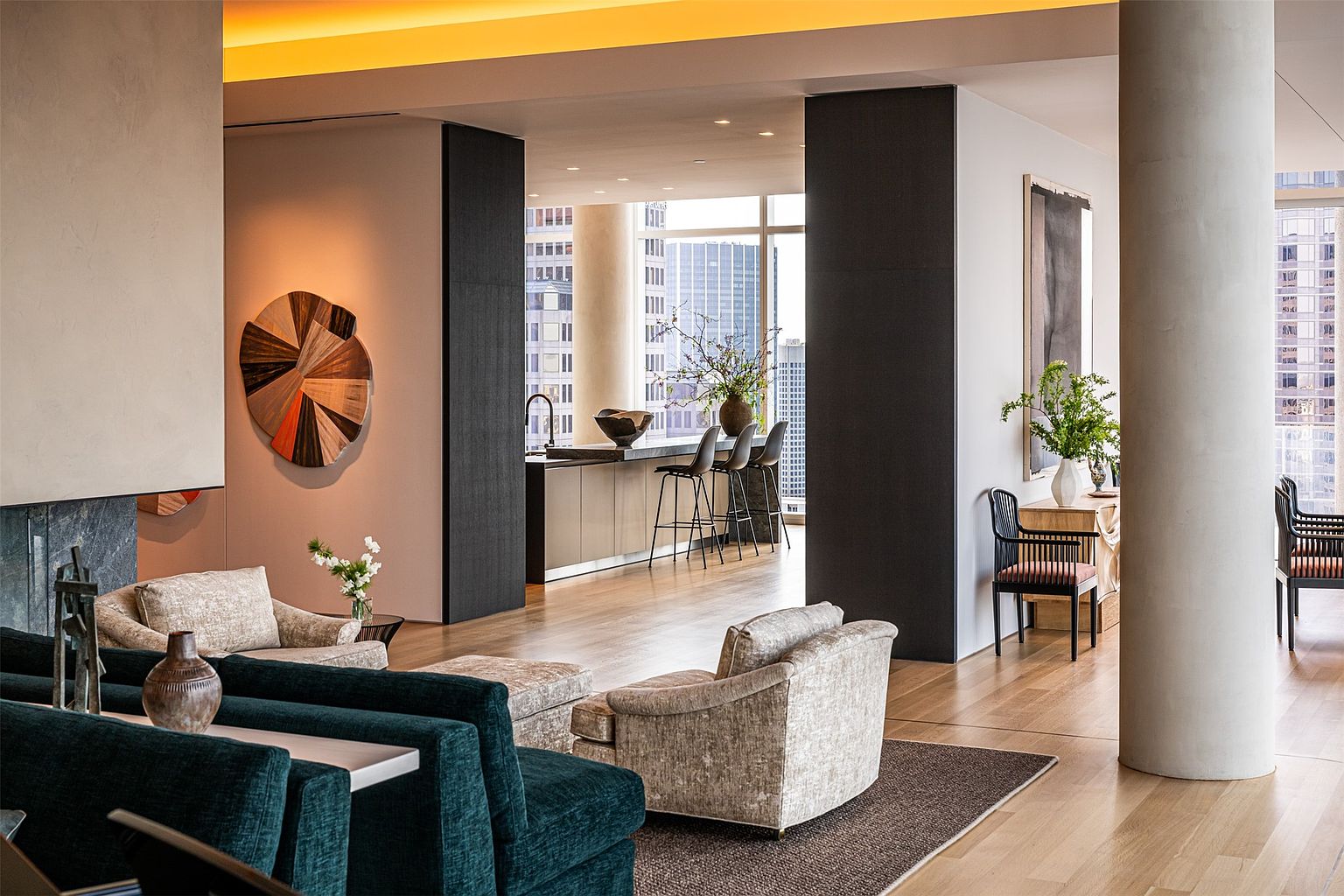 This sophisticated open-concept living space features a deep teal sofa in the foreground and plush, textured armchairs arranged on a dark area rug. The room flows seamlessly into a kitchen area with a breakfast bar and tall windows that offer expansive city views. The interior design is characterized by a neutral color palette, warm wood flooring, and curated art pieces, creating an atmosphere of refined urban luxury.