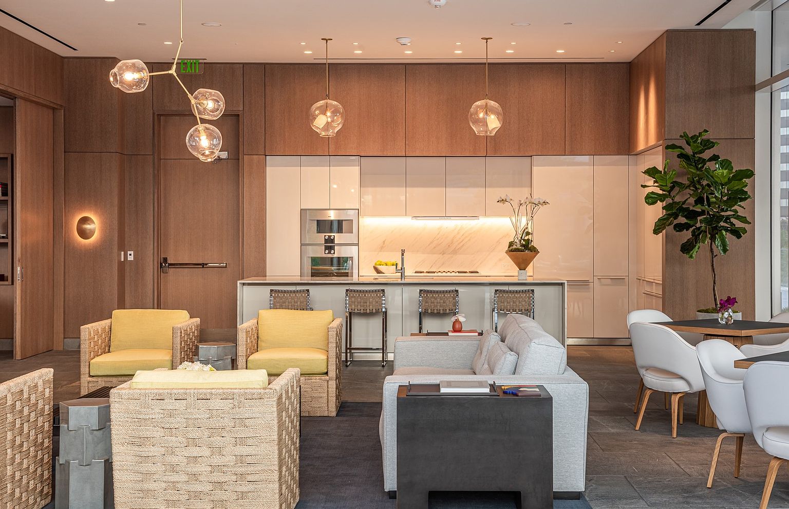 This sophisticated lounge area features a contemporary open-plan design with a sleek kitchen island, light wood cabinetry, and integrated stainless steel appliances. The space is furnished with comfortable seating, including yellow armchairs and a plush sofa, arranged on a dark rug to create a welcoming social environment. Large pendant lights hang above the island, while a tall indoor tree and modern dining area add to the upscale, professional aesthetic of this communal space.