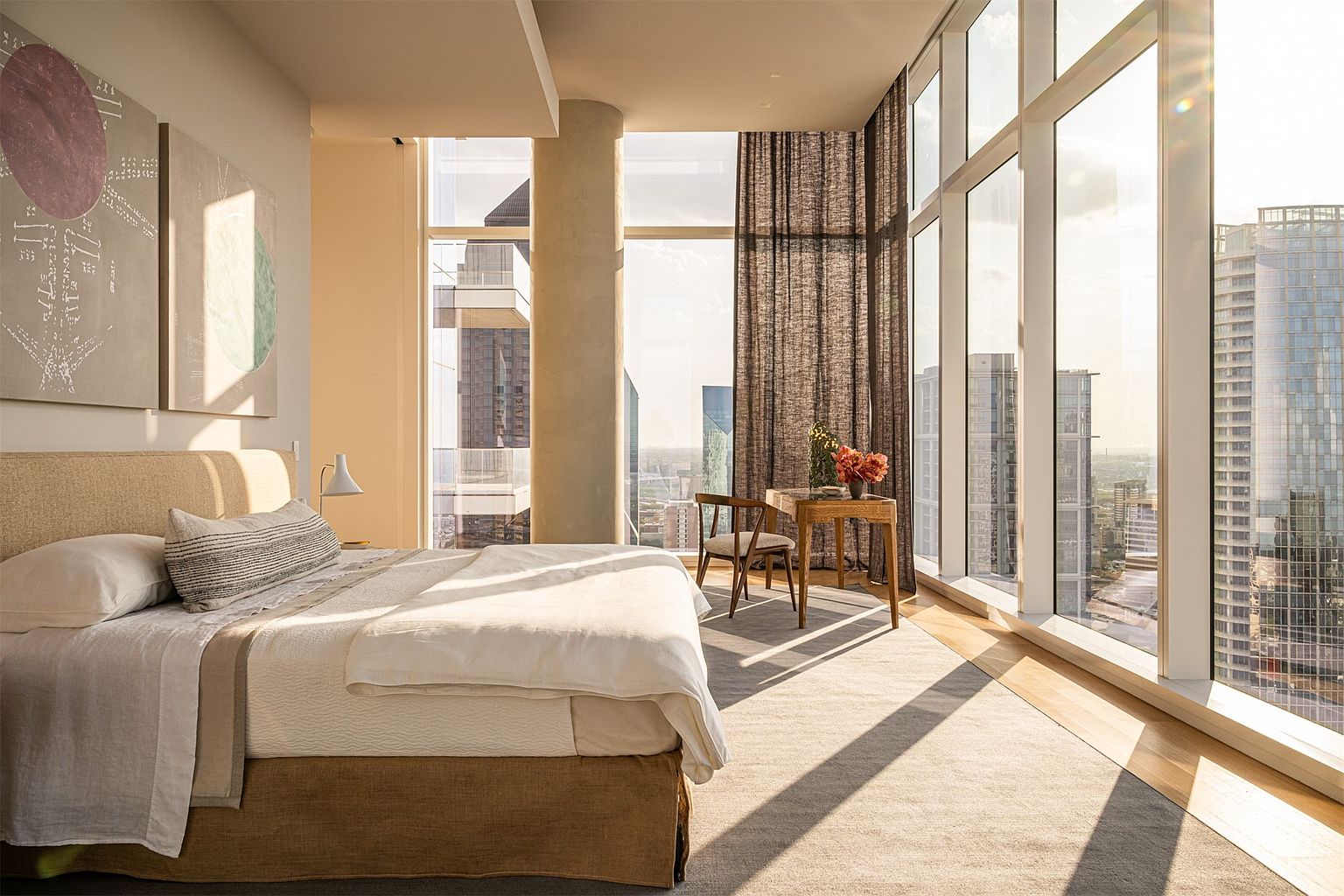 This sophisticated primary bedroom features expansive floor-to-ceiling windows that offer panoramic city views and bathe the space in warm, natural light. The room is anchored by a plush, neutral-toned bed and a minimalist wooden desk setup, creating a serene and luxurious atmosphere. A large area rug defines the space, while the clean lines and modern architectural elements emphasize a high-end, urban living experience.