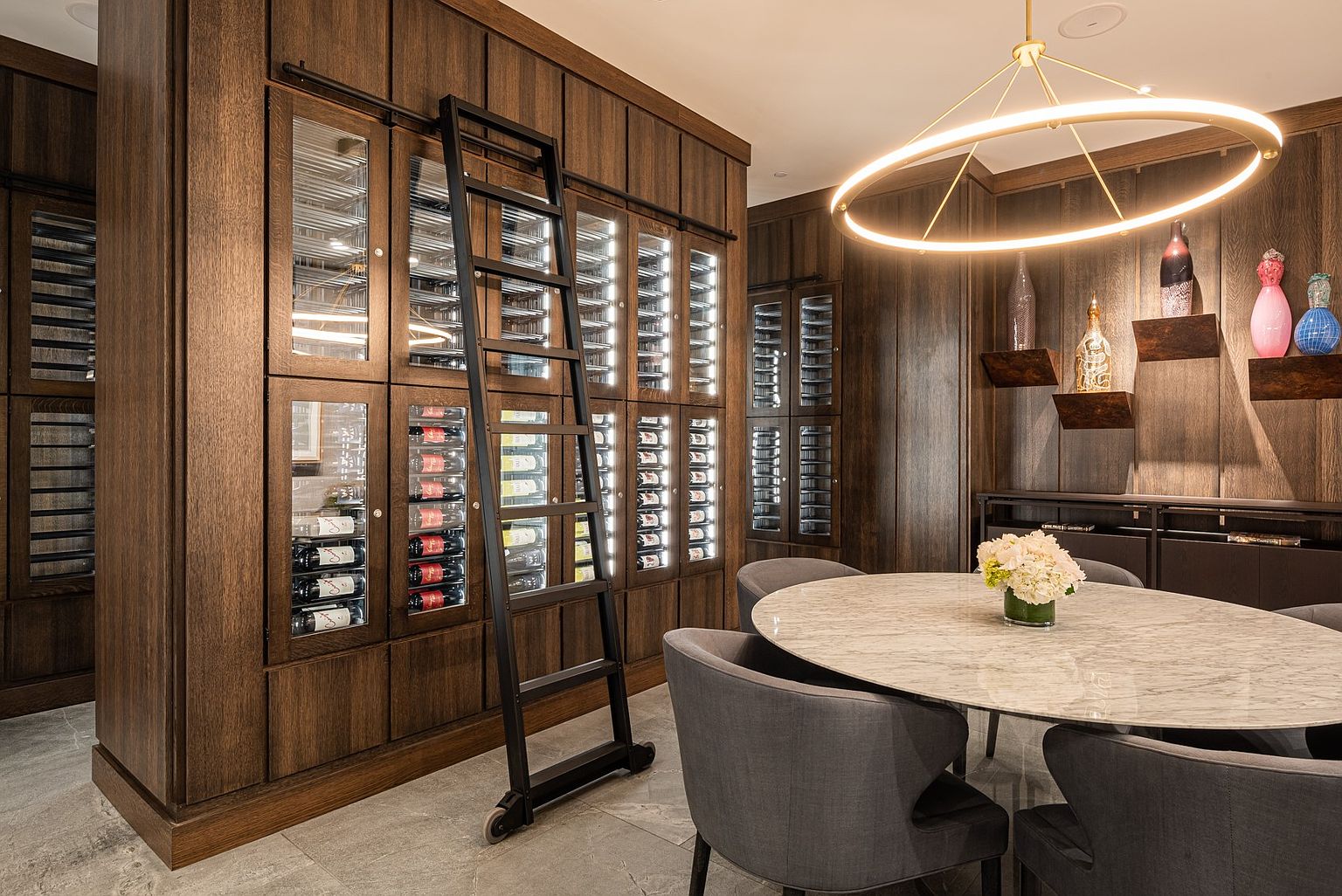 This sophisticated wine cellar features extensive custom wood cabinetry with integrated glass-front storage and internal lighting to showcase a wine collection. A rolling library-style ladder provides access to the upper shelves, while a central round stone-topped table surrounded by upholstered chairs creates an intimate tasting area. The room is illuminated by a large, modern circular pendant light, emphasizing the refined and luxurious atmosphere.