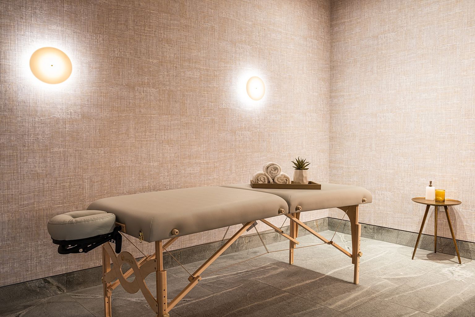 This serene spa treatment room features a professional massage table as the central focus, complete with a headrest and neatly rolled towels. The room is finished with textured, neutral-toned wallpaper and soft, warm wall-mounted lighting that creates a calming atmosphere. A small side table with candles and a potted succulent adds a touch of minimalist elegance to the space.