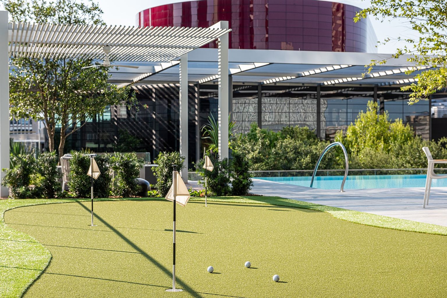 This outdoor amenity space features a putting green with three flag-marked holes and several golf balls scattered across the artificial turf. Adjacent to the green is a swimming pool with a curved metal handrail, set against a backdrop of lush greenery and a modern building with a red, cylindrical architectural feature. The area is shaded by a slatted pergola structure, creating a sophisticated and recreational atmosphere for residents.
