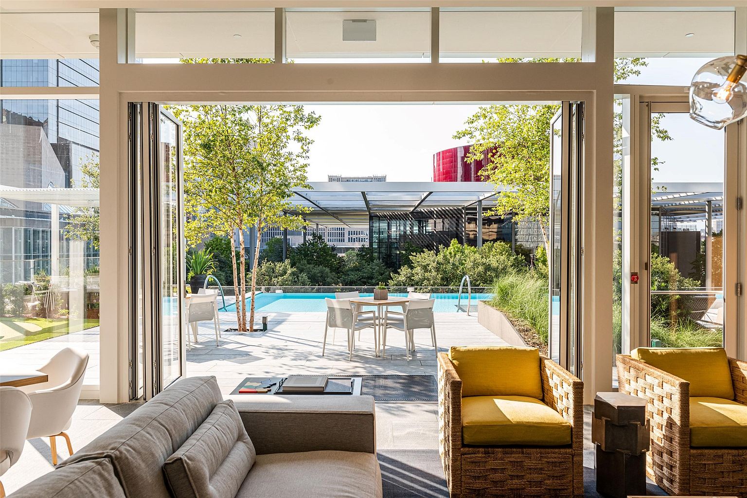 This sophisticated living space features a seamless transition to an outdoor terrace through large, folding glass doors. The interior is furnished with a plush grey sofa and two woven armchairs with yellow cushions, while the exterior patio offers a dining set and a clear view of a swimming pool surrounded by lush trees. The perspective from inside the room highlights the open-concept design and the bright, airy connection between the indoor lounge and the urban outdoor oasis.