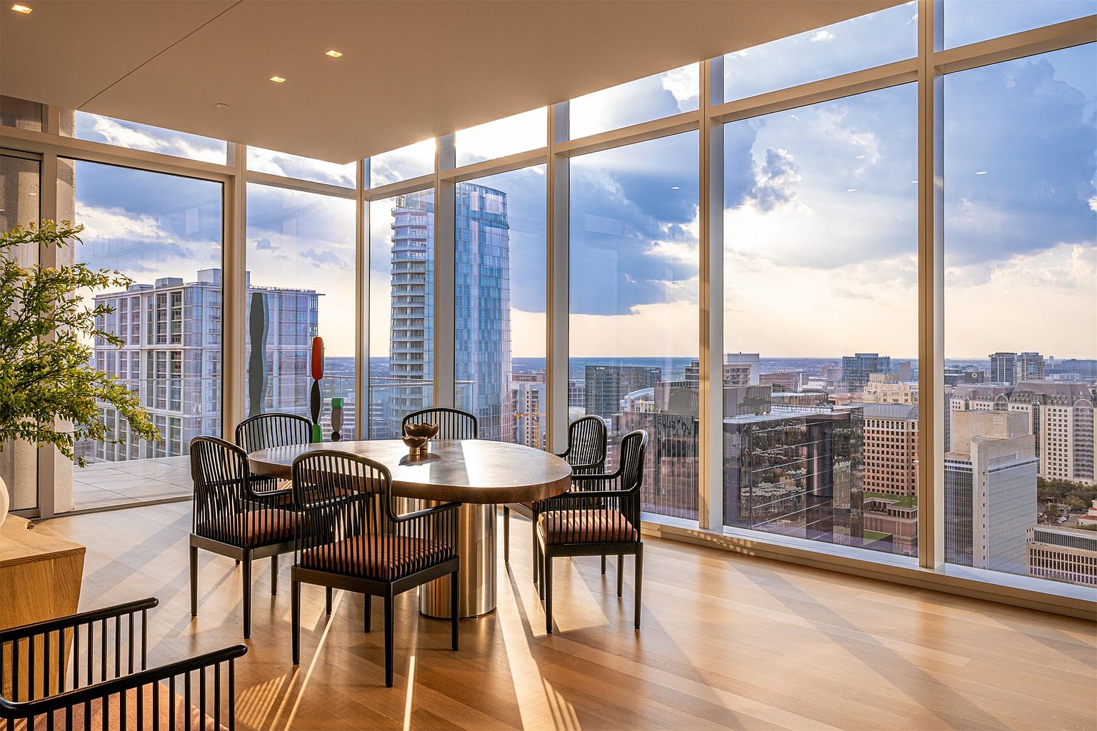 This sophisticated dining area features a round table surrounded by modern, slatted-back chairs, set against a backdrop of expansive floor-to-ceiling windows that offer a panoramic view of the city skyline. The room is bathed in warm, natural light, highlighting the light wood flooring and the minimalist, open-concept design. A sculptural art piece adds a touch of color to the space, creating an elegant and airy atmosphere perfect for high-rise urban living.