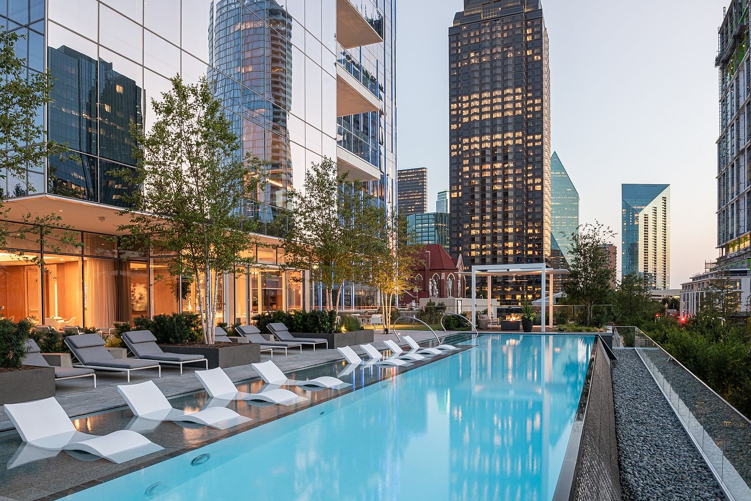 This sophisticated urban rooftop pool area features a long, rectangular infinity-edge pool lined with submerged lounge chairs. The deck is paved with stone and surrounded by lush landscaping, while the adjacent building showcases expansive glass walls that reflect the surrounding city skyline. The scene is captured at dusk, creating a serene and luxurious atmosphere with warm interior lighting glowing from the building's common areas.
