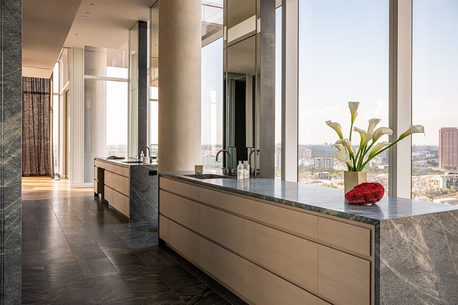 This sophisticated primary bathroom features dual floating vanities with light wood cabinetry and dark stone countertops, set against a backdrop of expansive floor-to-ceiling windows that offer panoramic city views. The space is anchored by dark stone flooring and a large structural column, creating a sleek, high-end aesthetic. A vase of calla lilies and a decorative red coral piece add a touch of organic elegance to the minimalist, modern design.