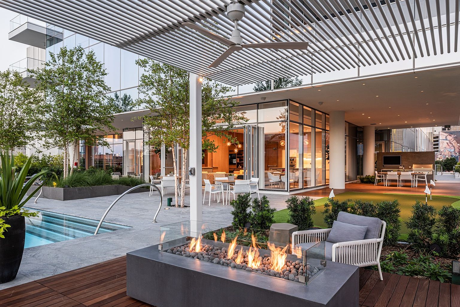This sophisticated outdoor amenity space features a modern fire pit with a glass surround, set on a wooden deck overlooking a swimming pool and a putting green. The area is sheltered by a sleek, slatted metal pergola and connects seamlessly to an indoor lounge through large glass walls. The overall aesthetic is contemporary and luxurious, designed for high-end social gatherings and relaxation.