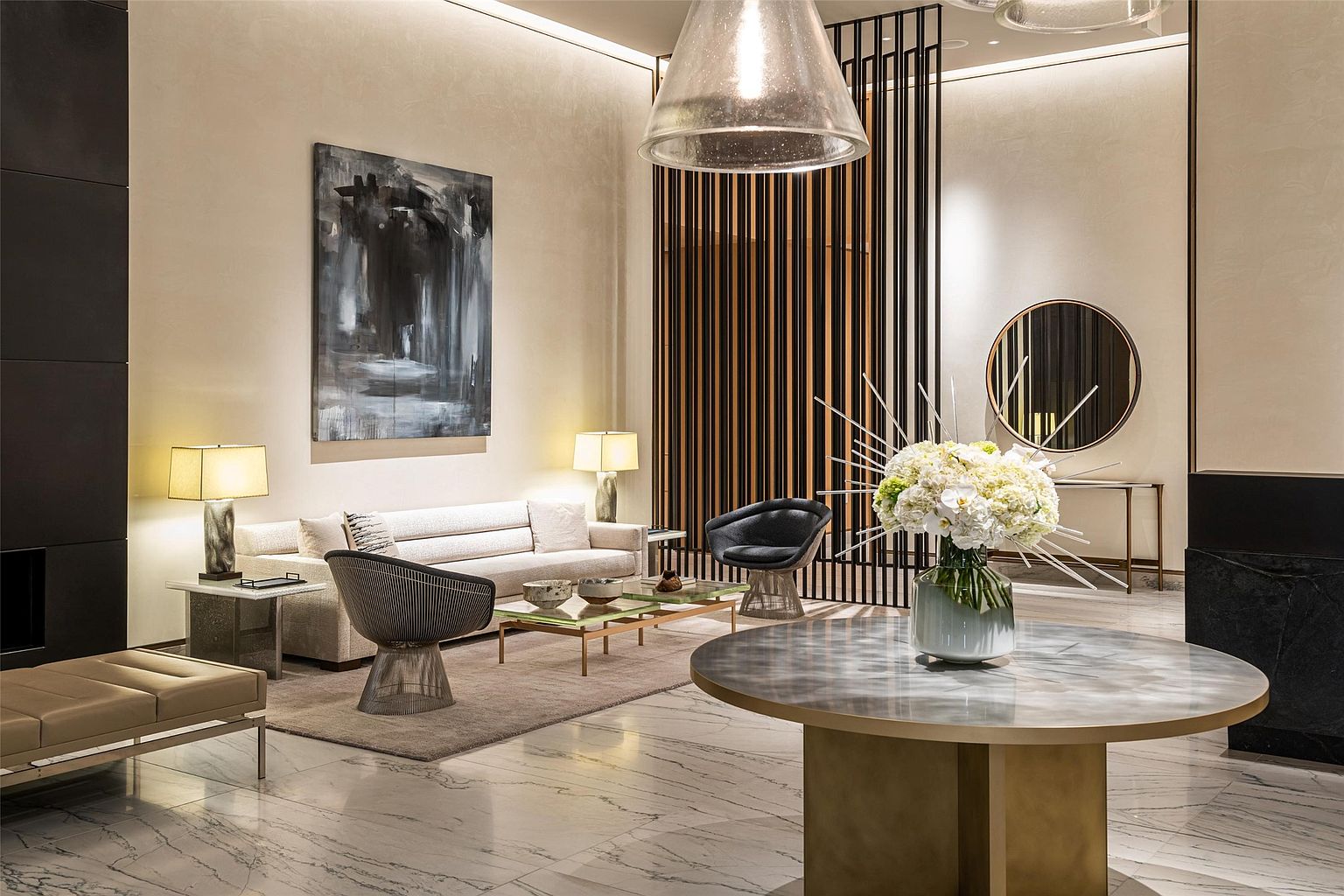 This sophisticated lobby or lounge area features a high-end, contemporary aesthetic with polished stone flooring and a neutral color palette. A central circular table with a stone top serves as a focal point, complemented by a seating area with a plush sofa, modern wire-frame chairs, and a large abstract painting. The space is defined by a dramatic vertical slat screen and a large, textured glass pendant light, creating an atmosphere of refined luxury.