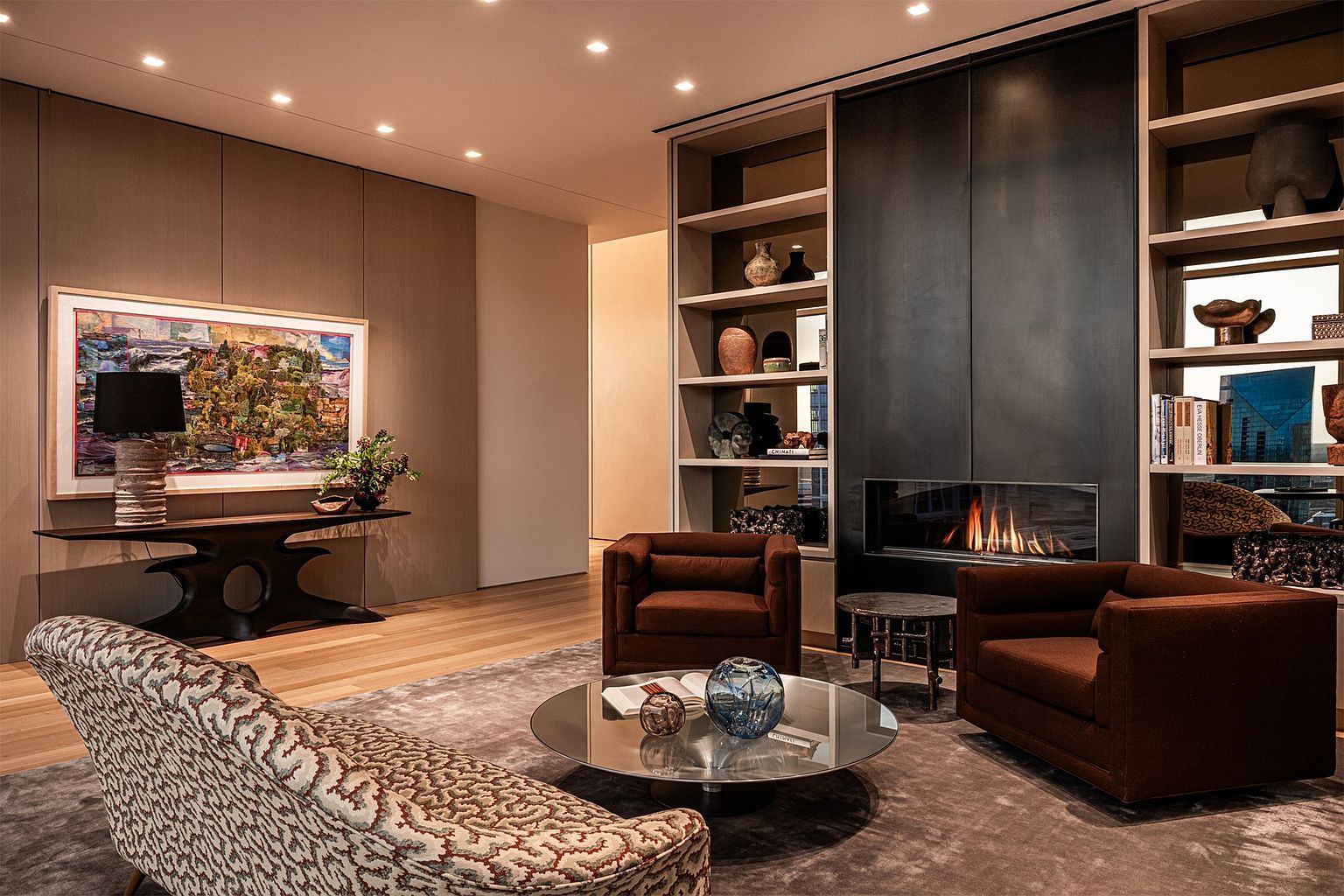 This sophisticated living room features a warm, neutral color palette with a prominent dark metal fireplace surround integrated into a custom shelving unit. The space is furnished with two plush brown armchairs and a patterned sofa, centered around a circular glass coffee table on a soft grey rug. A console table with a sculptural base and a large, vibrant piece of abstract art adds a refined, gallery-like aesthetic to the room.