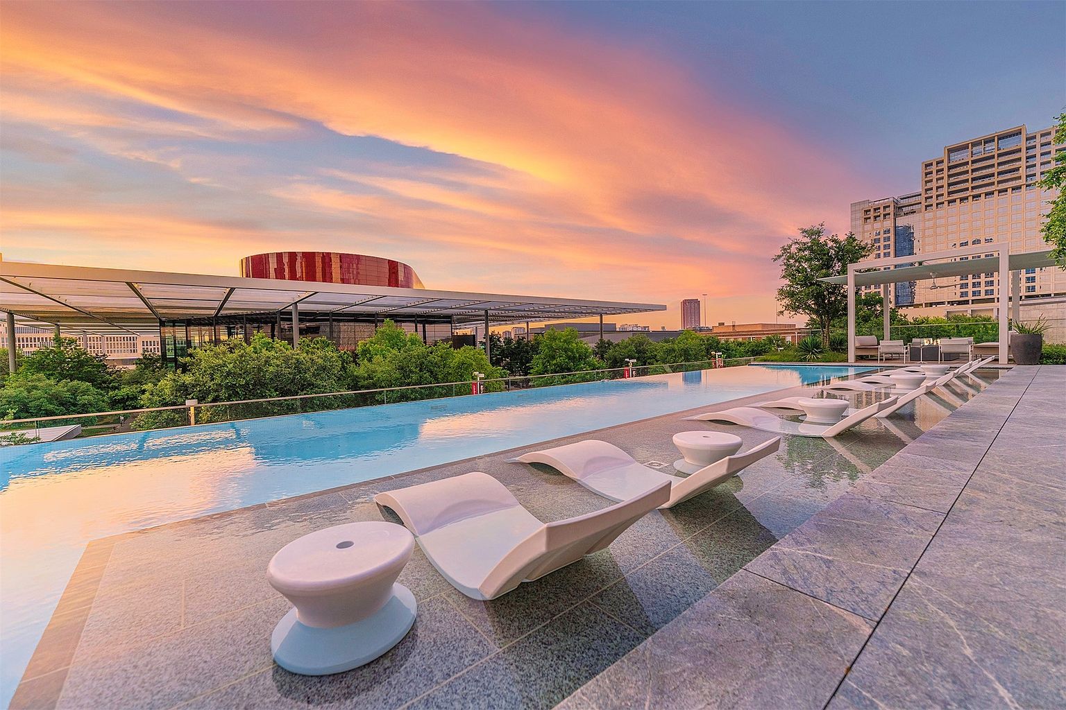 This stunning rooftop infinity-edge pool offers a luxurious urban retreat with panoramic views of the city skyline at sunset. The pool deck is lined with sleek, white lounge chairs and matching side tables, set against a backdrop of lush green trees and modern architectural structures. The warm, vibrant colors of the evening sky reflect beautifully on the water's surface, creating an inviting and sophisticated atmosphere.