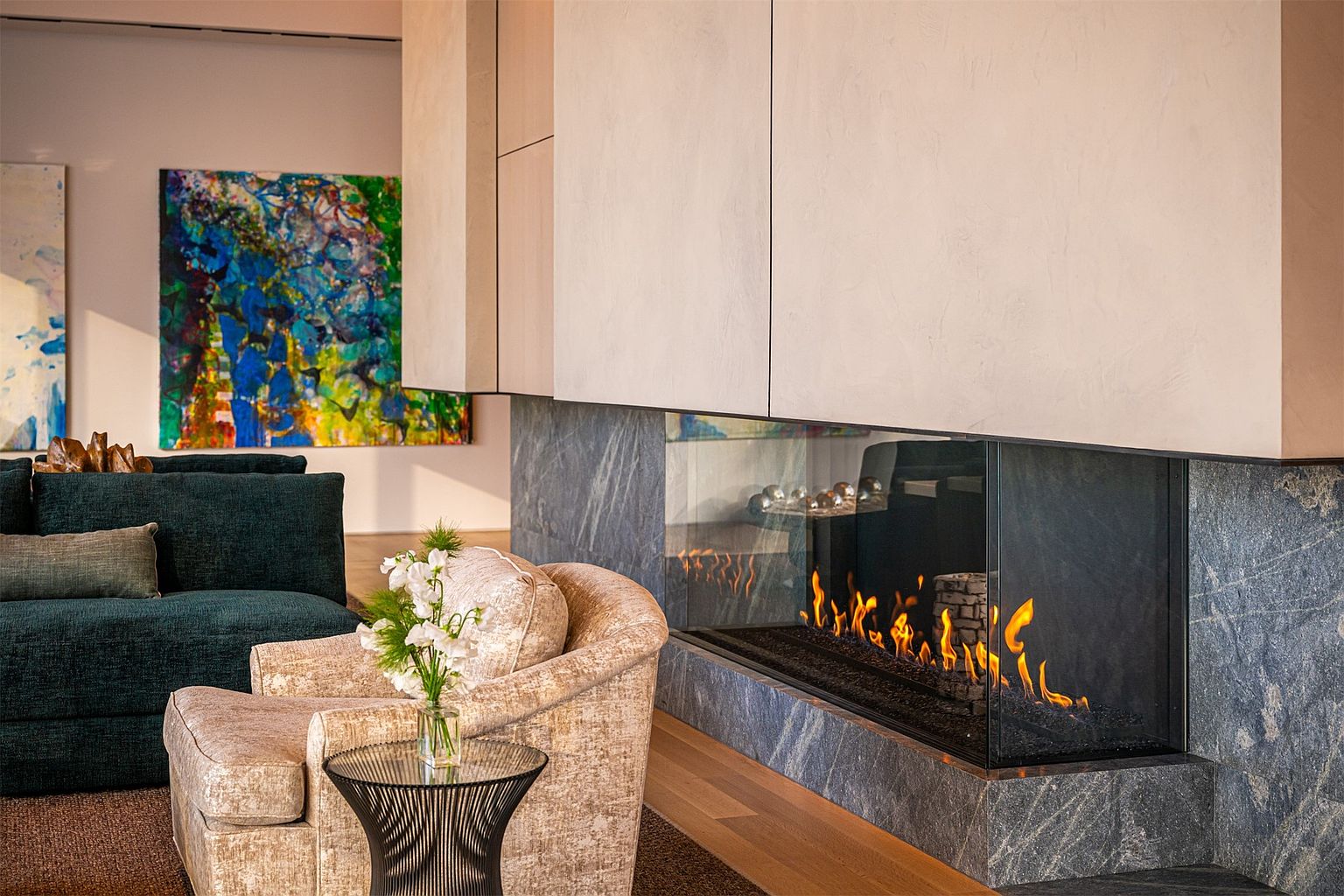This sophisticated living space features a prominent, multi-sided fireplace with a dark stone base and a tall, light-toned plaster surround. A plush, textured armchair sits beside a small glass-topped side table with a floral arrangement, while a deep-toned sofa is visible in the background. The room is accented by a vibrant, colorful abstract painting on the wall, creating a refined and artistic atmosphere.