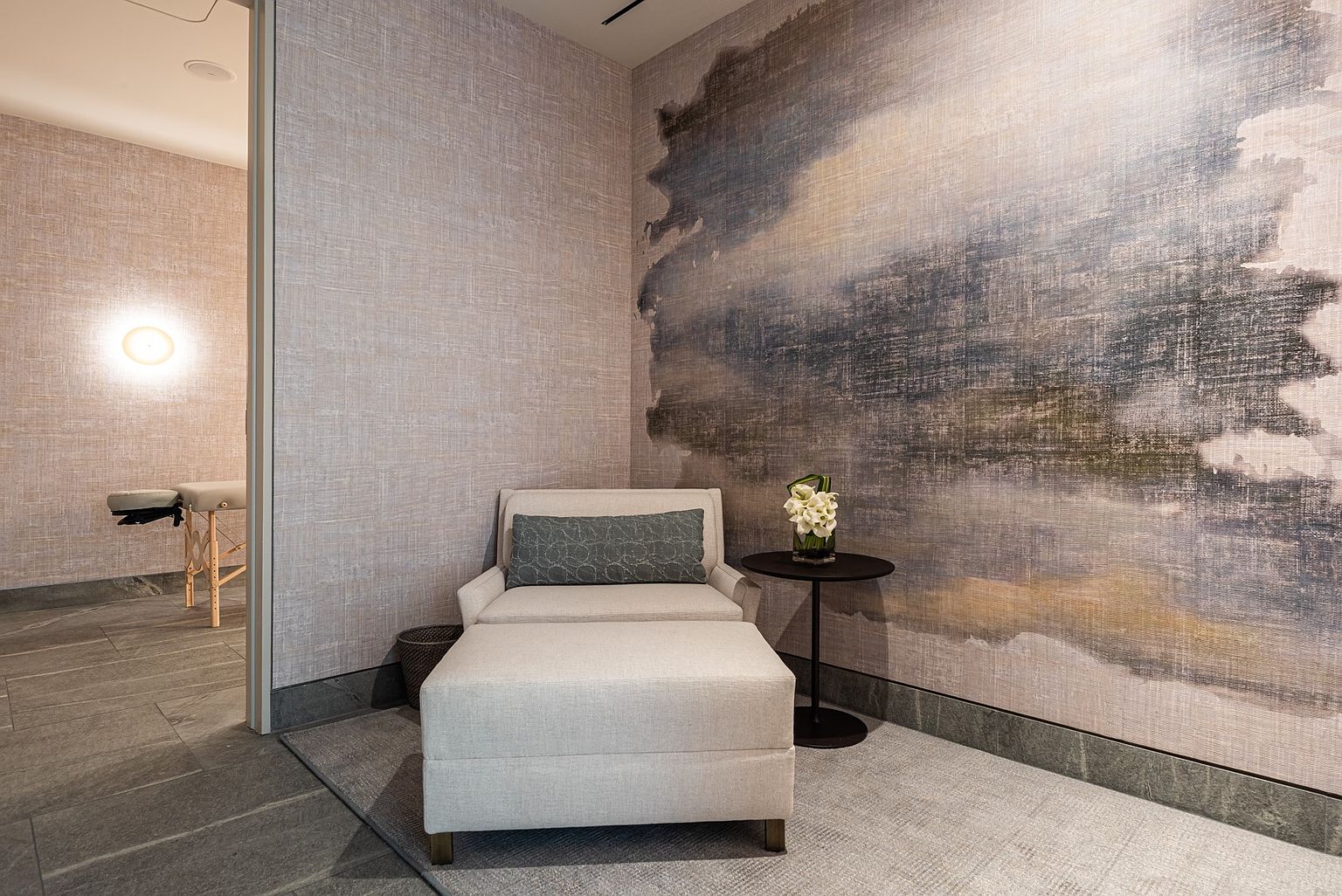 This serene relaxation area features a plush lounge chair with an ottoman, accented by a patterned bolster pillow and a small side table holding a floral arrangement. The space is defined by a large-scale, abstract mural on the wall and textured wall coverings, creating a calming atmosphere. A doorway leads into an adjacent treatment room, which contains a massage table and wall-mounted lighting, suggesting a high-end spa or wellness facility.