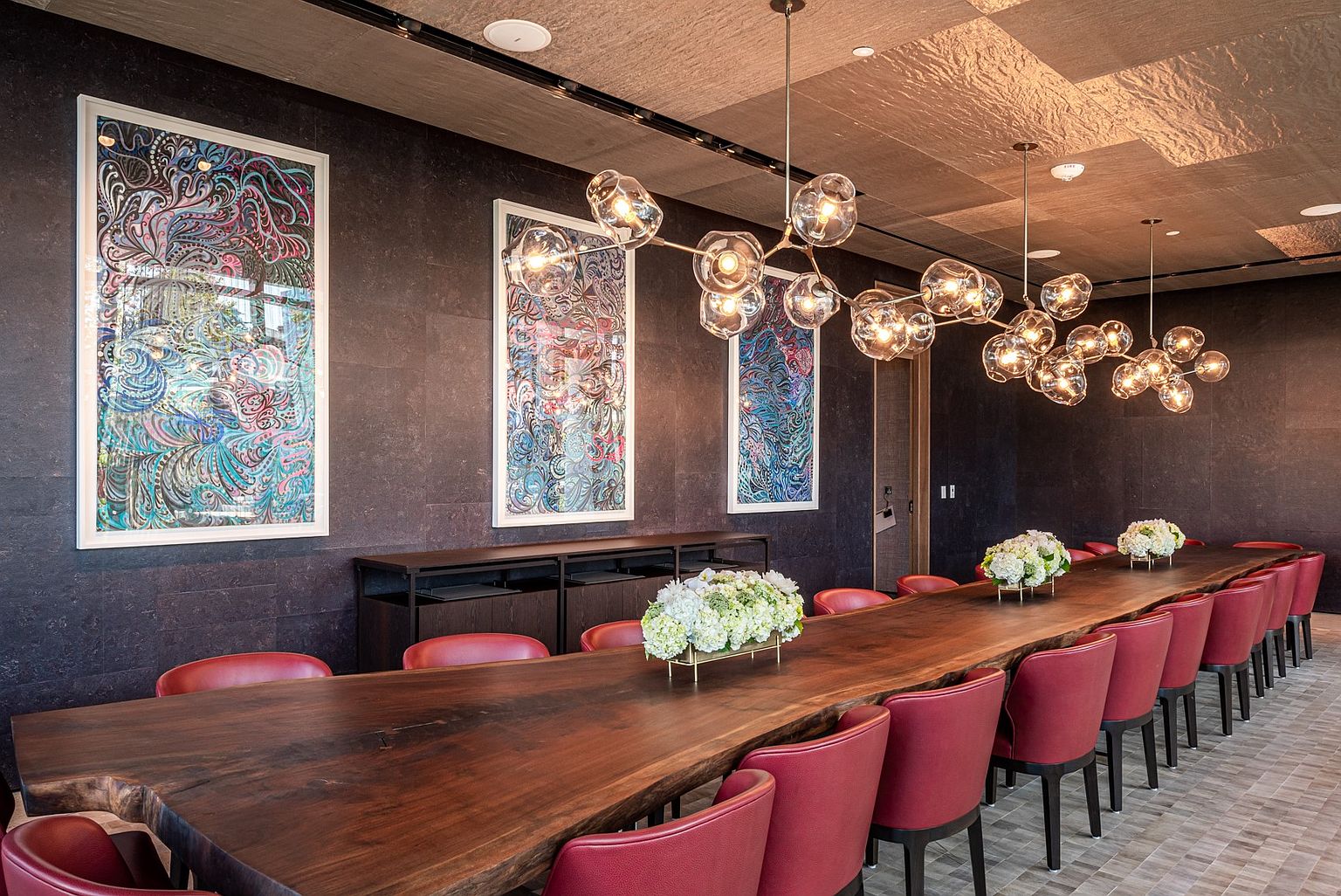 This sophisticated dining room features a long, live-edge wood table surrounded by vibrant red chairs, creating a bold and elegant focal point. The dark, textured wall serves as a dramatic backdrop for three large, colorful abstract art pieces, while a series of modern, multi-globe pendant lights hang above the table. The space is further accented by floral centerpieces and a unique, textured ceiling, offering a luxurious and contemporary atmosphere suitable for formal gatherings.