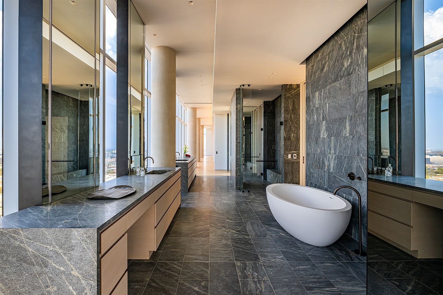 This luxurious primary bathroom features a sleek, modern design with dark stone surfaces and light wood cabinetry. A freestanding white soaking tub serves as a focal point, complemented by a long vanity and floor-to-ceiling windows that offer expansive city views. The space is characterized by its clean lines, high-end finishes, and a bright, airy atmosphere created by the natural light.