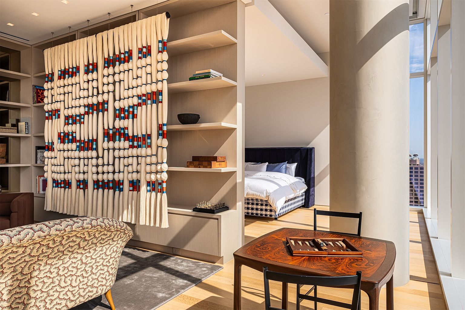 This sophisticated interior space features an open-concept layout that seamlessly connects a living area with a bedroom nook. A prominent, large-scale fiber art installation serves as a room divider, positioned next to custom light wood shelving. In the foreground, a dark wood game table with a backgammon set sits near floor-to-ceiling windows that offer expansive views, while a patterned armchair adds a touch of texture to the room.