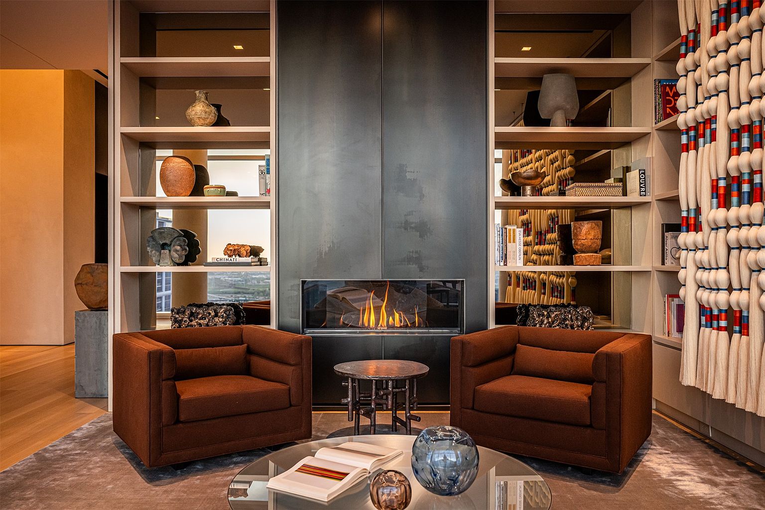 This sophisticated living area features a central fireplace with a dark, full-height metal surround flanked by symmetrical built-in shelving units. Two plush, brown armchairs are positioned symmetrically in front of the fireplace, centered around a circular glass coffee table. The room exudes a refined, contemporary aesthetic with curated decor, including textured wall art and sculptural objects, creating a warm and inviting atmosphere.