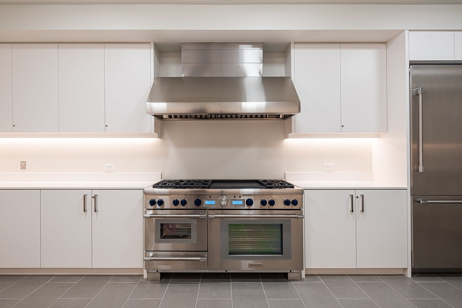 This modern kitchen features a symmetrical layout with crisp white cabinetry and a professional-grade stainless steel range as the central focal point. A sleek stainless steel range hood is mounted above the stove, complemented by under-cabinet lighting that illuminates the clean, neutral backsplash. The space is finished with large-format grey floor tiles and a full-sized stainless steel refrigerator integrated into the cabinetry wall, creating a polished and functional culinary environment.