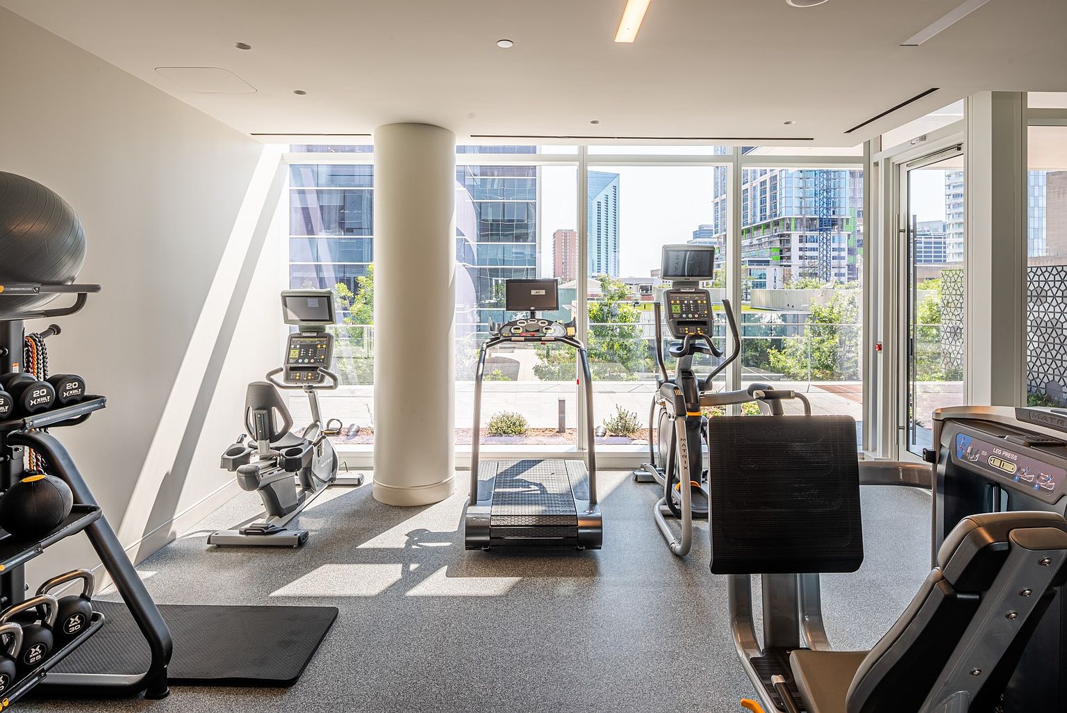 This bright and modern home gym features a variety of professional-grade fitness equipment, including a treadmill, elliptical, and leg press machine. The space is bathed in natural light from large windows that offer an expansive view of the surrounding urban cityscape. The room has a clean, minimalist aesthetic with neutral flooring and a structural column, creating an inviting atmosphere for a workout.