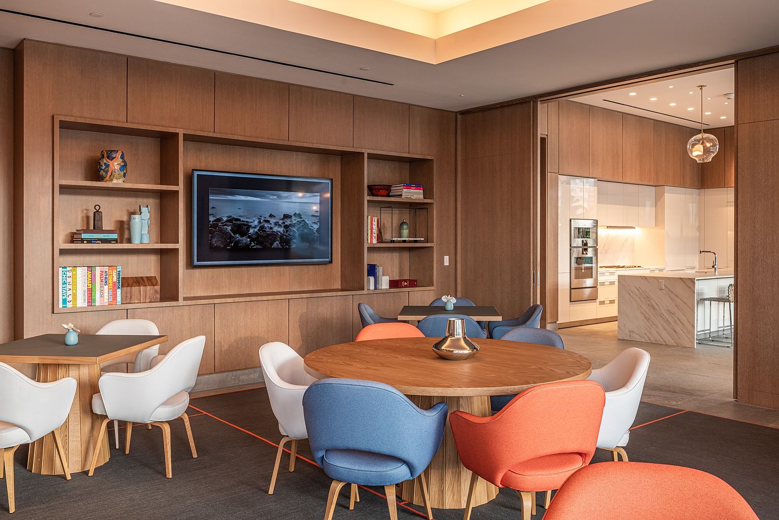 This sophisticated lounge area features a warm, wood-paneled wall with built-in shelving and a central television, creating a cozy yet modern atmosphere. The space is furnished with multiple round tables paired with colorful, mid-century modern style chairs in shades of blue and orange. Through an open doorway, a glimpse of a sleek, contemporary kitchen with a stone-topped island and stainless steel appliances suggests a high-end residential amenity space.