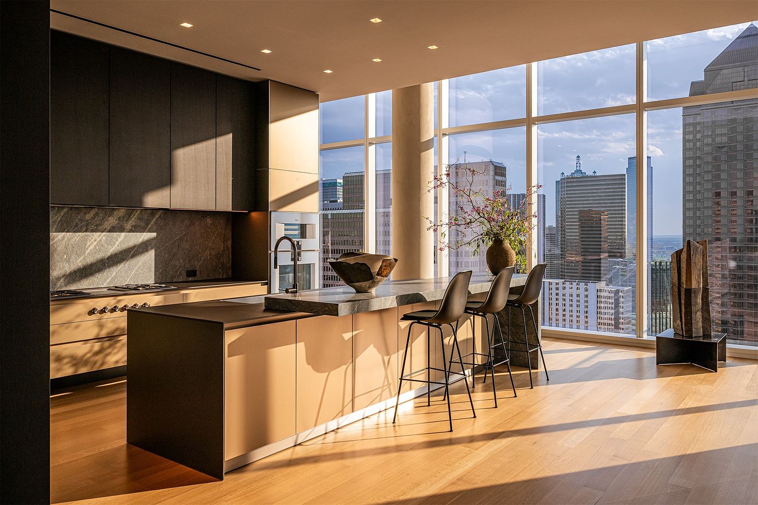 This sophisticated, modern kitchen features a sleek island with a stone countertop and three minimalist bar stools, set against a backdrop of expansive floor-to-ceiling windows that offer a panoramic view of the city skyline. The space is defined by a clean, high-contrast aesthetic, combining dark cabinetry with light wood-toned lower cabinets and warm, natural light that highlights the wood flooring. A sculptural bowl sits on the island, while a tall, artistic vase arrangement adds a touch of organic elegance to the bright, airy atmosphere.