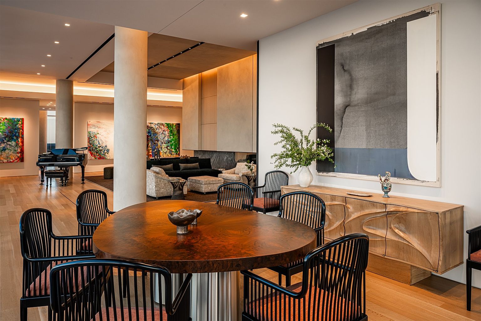 This sophisticated dining area features a large, rounded wooden table surrounded by black spindle-back chairs with upholstered seats. The space is anchored by a unique, textured wooden sideboard and a large abstract painting, creating a gallery-like atmosphere. In the background, an open-concept living area with a grand piano and plush seating is visible, showcasing an expansive, high-end interior design with warm lighting and neutral tones.