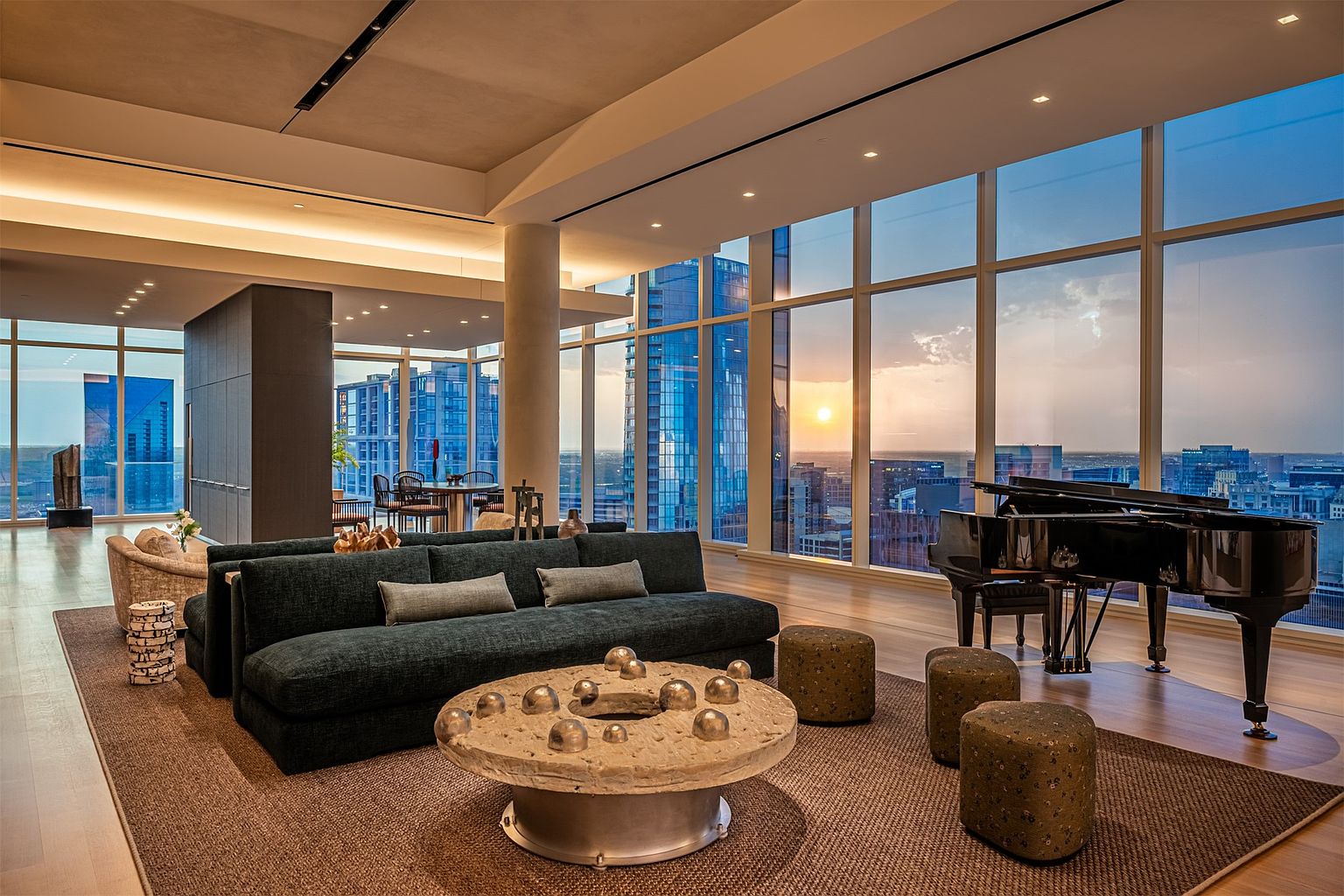 This expansive, high-rise living room features a sophisticated open-concept layout with floor-to-ceiling windows that frame a panoramic sunset view of the city skyline. The space is anchored by a dark, plush sofa and a unique, circular stone coffee table adorned with metallic spheres, complemented by several textured ottomans. A grand piano sits elegantly near the glass, creating a refined atmosphere that blends modern luxury with artistic flair.