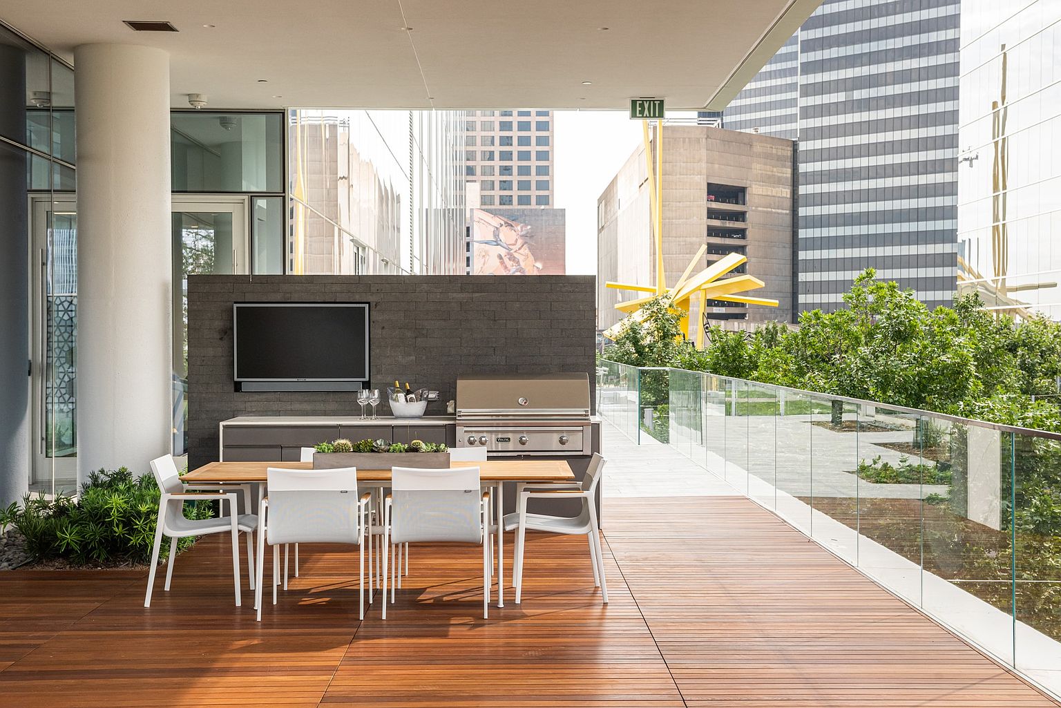 This modern, covered outdoor terrace features high-end amenities including a built-in stainless steel grill and a wall-mounted television set against a dark, textured stone feature wall. A sleek wooden dining table with four white chairs sits on the warm-toned wood decking, which is bordered by a minimalist glass railing. The space offers an urban perspective, overlooking a landscaped garden area and surrounding city skyscrapers.