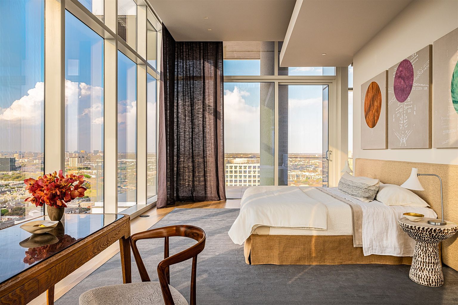 This sophisticated primary bedroom features expansive floor-to-ceiling windows that offer a panoramic city view and bathe the space in warm, natural light. The room is furnished with a plush bed, a unique patterned side table, and a sleek wooden desk paired with a curved chair. Abstract circular artwork adorns the wall above the bed, contributing to the modern, high-rise aesthetic of the suite.