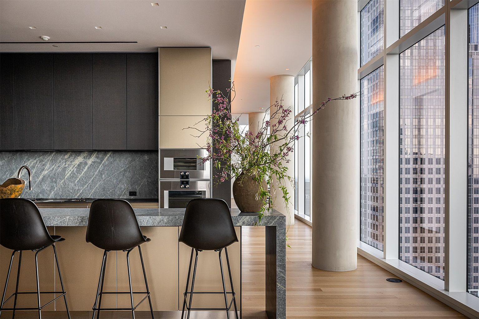 This sophisticated, modern kitchen features a sleek island with a stone countertop and three dark stools, set against a backdrop of dark cabinetry and a stone backsplash. A large, organic-shaped vase filled with flowering branches serves as a striking centerpiece on the island, while floor-to-ceiling windows provide an expansive view of the city skyline. The space is defined by its clean lines, neutral color palette, and a prominent structural column, creating an atmosphere of refined urban luxury.