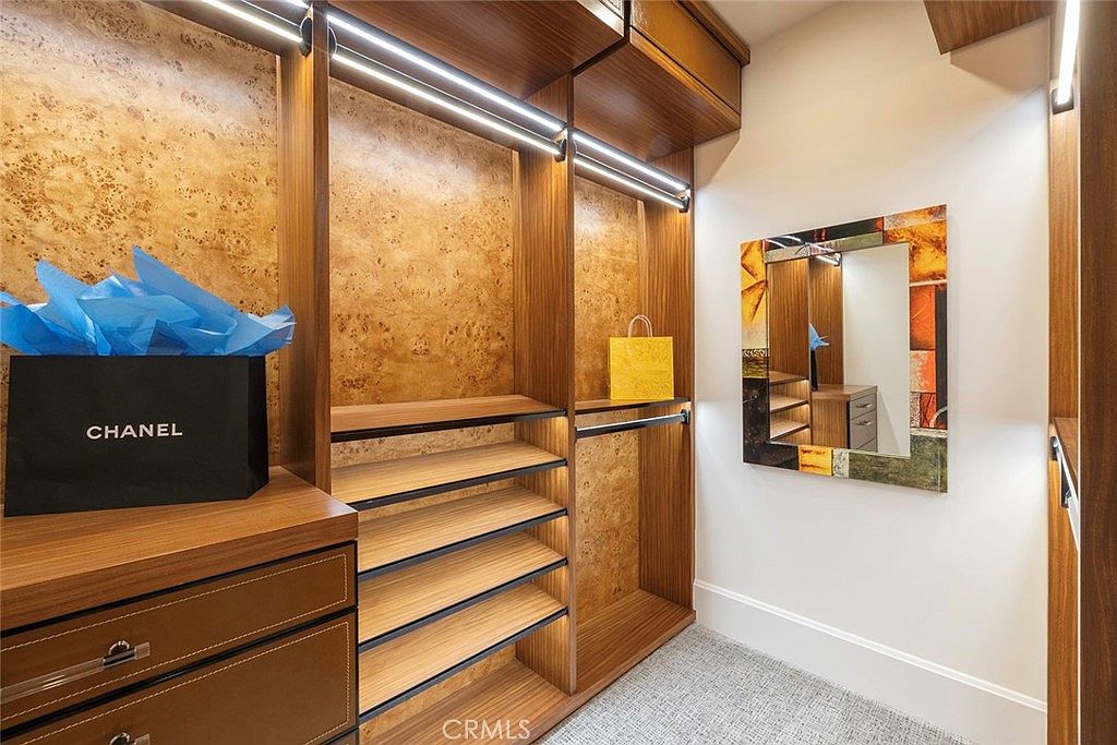 This is a well-organized walk-in closet featuring custom wood cabinetry with built-in shelving and drawers. The closet is illuminated by modern strip lighting, and a decorative mirror adds a touch of elegance. A Chanel shopping bag and a yellow handbag are placed on the shelves, suggesting a high-end lifestyle.
