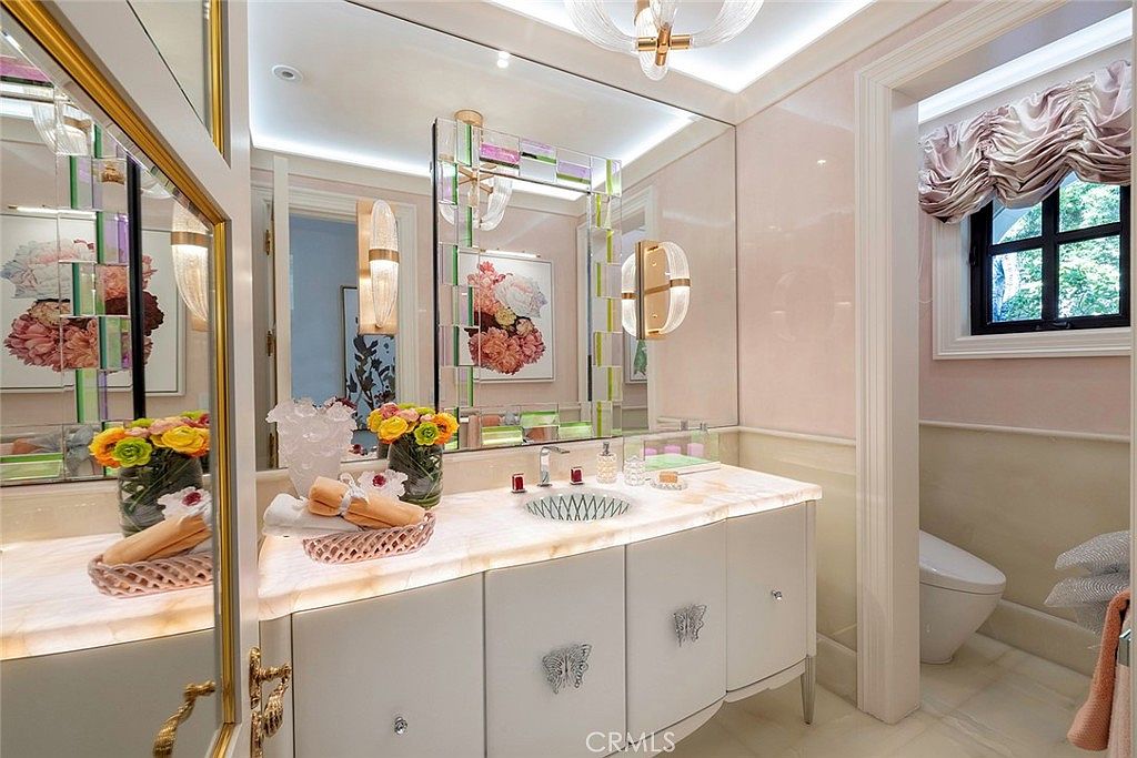 This is a luxurious bathroom featuring a custom vanity with a marble countertop and decorative butterfly hardware. The room has a pink color scheme, with a large mirror and unique lighting fixtures. A glimpse into an adjacent toilet room reveals a window with elegant drapery, adding to the overall opulent and well-designed aesthetic.