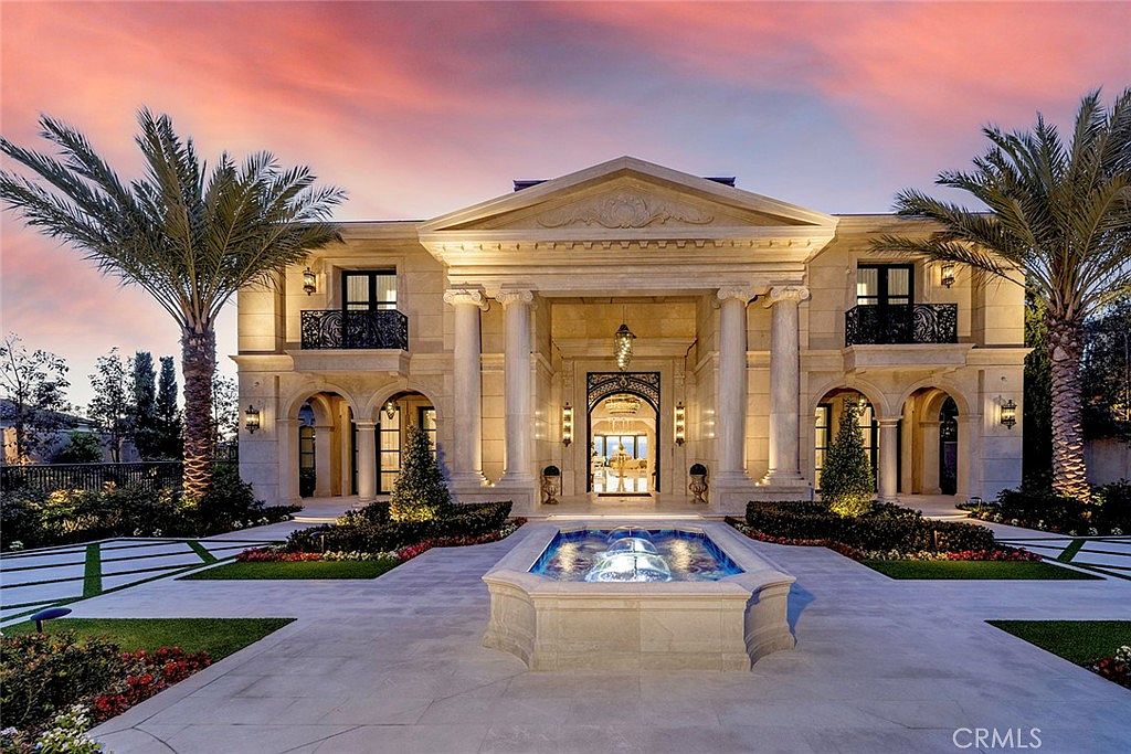 This is a stunning front view of a grand estate, showcasing its impressive architectural design with prominent columns, arched entryways, and symmetrical layout. The property features meticulously landscaped grounds, including palm trees and a central water fountain, enhancing its luxurious appeal. The warm lighting and picturesque sunset create an inviting and elegant atmosphere.