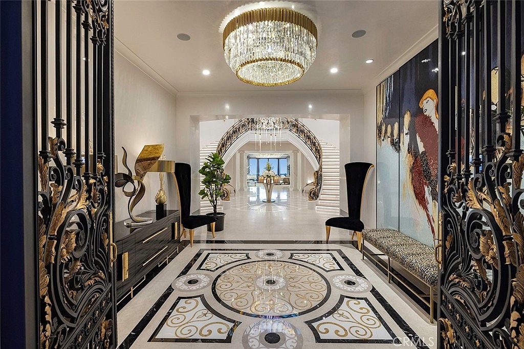 This grand foyer features an ornate black and gold gate leading into a luxurious space with a striking patterned marble floor, a large crystal chandelier, and a sweeping staircase. The hallway is decorated with high-end furnishings, including tall black chairs and a modern art piece, creating an opulent and inviting atmosphere. The perspective is from the entrance, looking into the grand hallway.