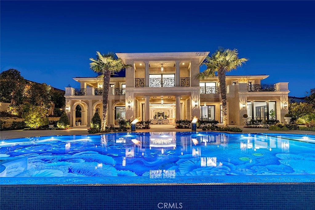 This image showcases a luxurious estate with a stunning pool as its focal point. The pool features intricate blue tile designs and reflects the elegant architecture of the home, which includes balconies, columns, and palm trees. The scene is set at dusk, with the home's lighting creating a warm and inviting ambiance.