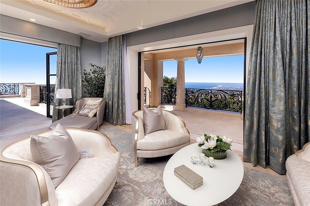 This elegant living room features luxurious seating arrangements with plush armchairs and a chaise lounge, complemented by a round coffee table adorned with floral arrangements. The room opens onto a spacious balcony with wrought iron railings and stunning ocean views, creating a seamless indoor-outdoor living experience. The interior is decorated with sophisticated curtains and a neutral color palette, enhancing the room's serene and upscale ambiance.