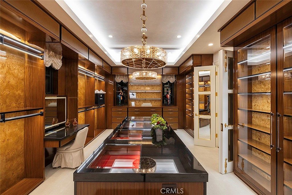 This is a luxurious walk-in closet featuring custom wood cabinetry with glass doors and integrated lighting. A large central island with a black countertop provides ample space for organization and display. The closet is illuminated by a decorative chandelier, adding a touch of elegance to the space.