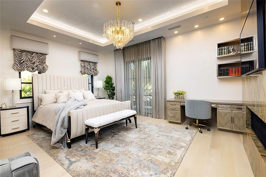 This is a luxurious primary bedroom featuring a large upholstered bed with a tufted headboard, complemented by a bench at the foot of the bed. The room is decorated in neutral tones with a patterned rug, elegant window treatments, and a crystal chandelier, creating a sophisticated and serene atmosphere. A desk area with built-in shelving and a TV completes the space.